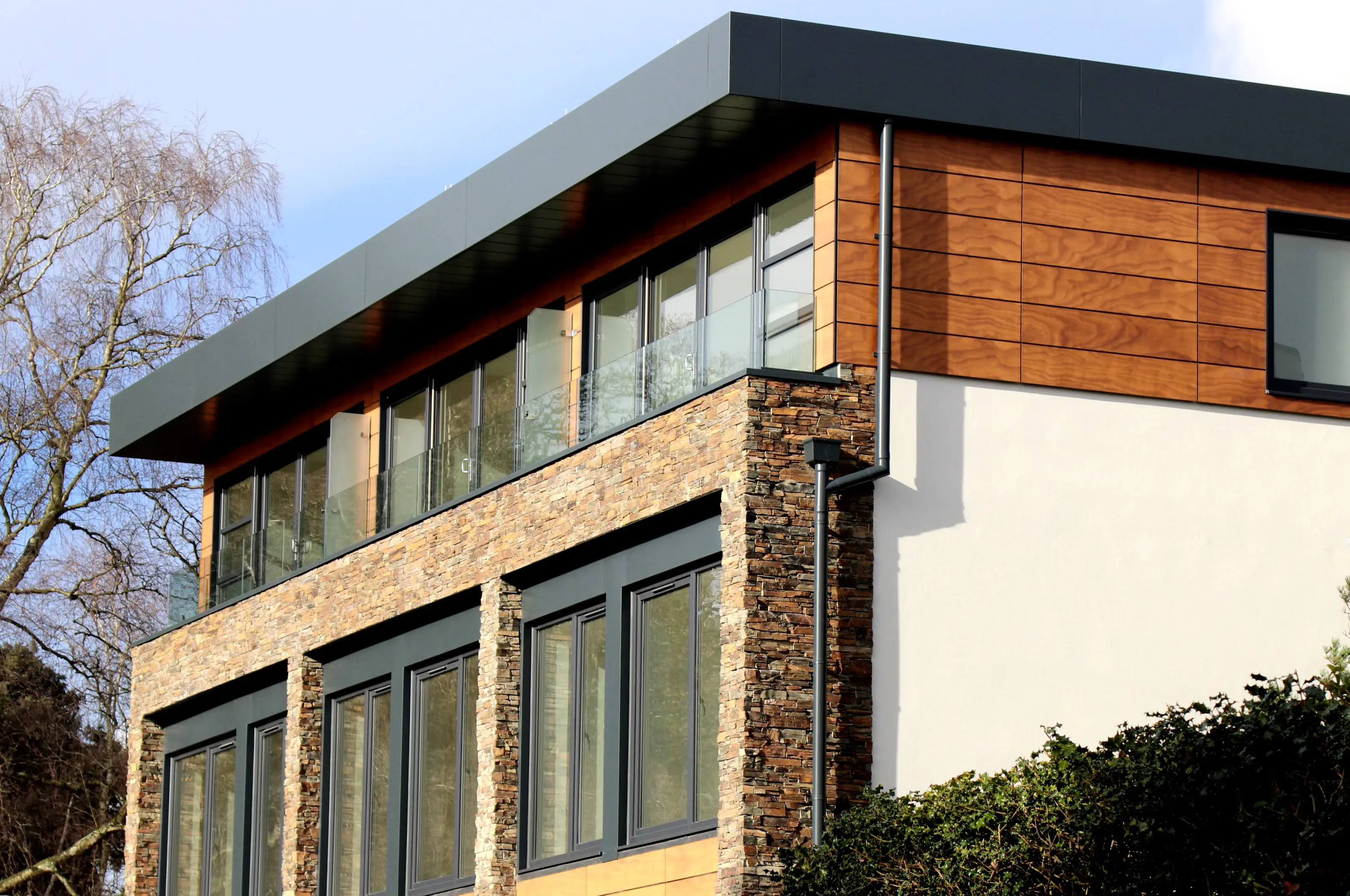 Modern residential façade showing wood cladding, stonework, windows, roof edges, and drainage components
